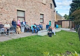 Photo showing approximately 12 people - plus one dog - sitting or standing outside of Ruspidge Memorial Hall in the new community garden.