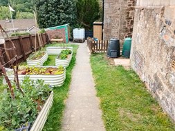 Photo showing part of the garden at Ruspidge Memorial Hall. The photo shows a path, two compost bins, a grassy area and some raised beds which are planted with a variety of plants and vegetables.